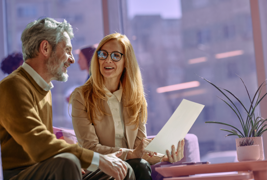 Two employees are talking about an HR policy change while seated on an office couch.