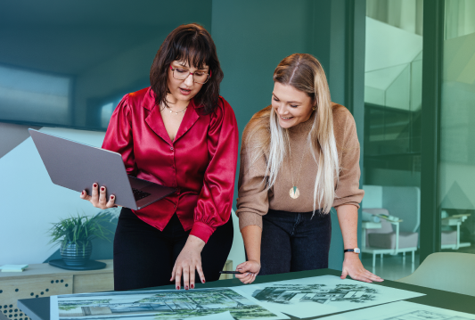 Two employees are standing at a table, working on project paperwork together while smiling?