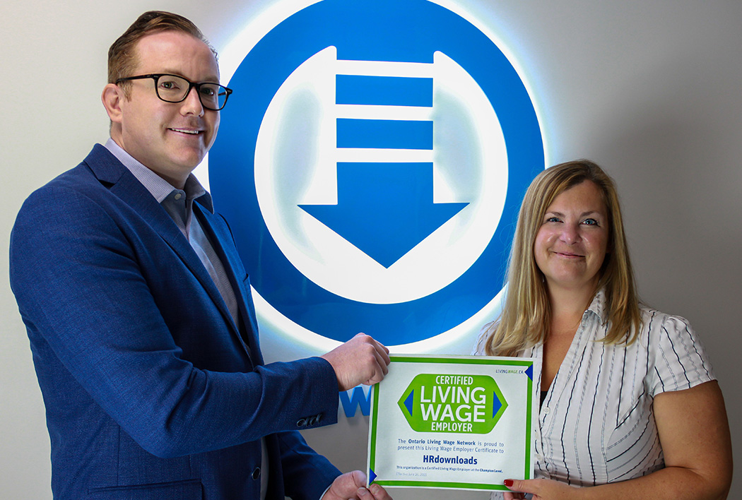 Neil Poutney and Sharon Bunce standing in front of Citation Canada, formerly HRdownloads, logo, holding the Living Wage Certified Certificate