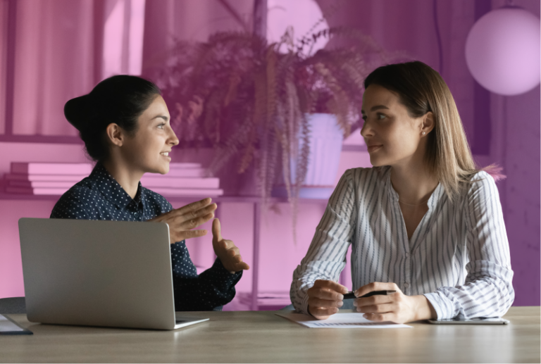 two employees are sitting next to each other at a desk with one laptop conversing