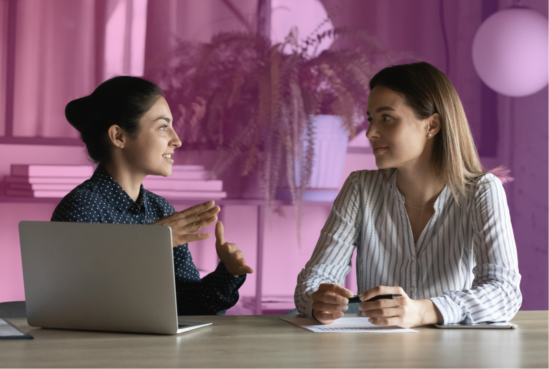 two employees are sitting next to each other at a desk with one laptop conversing