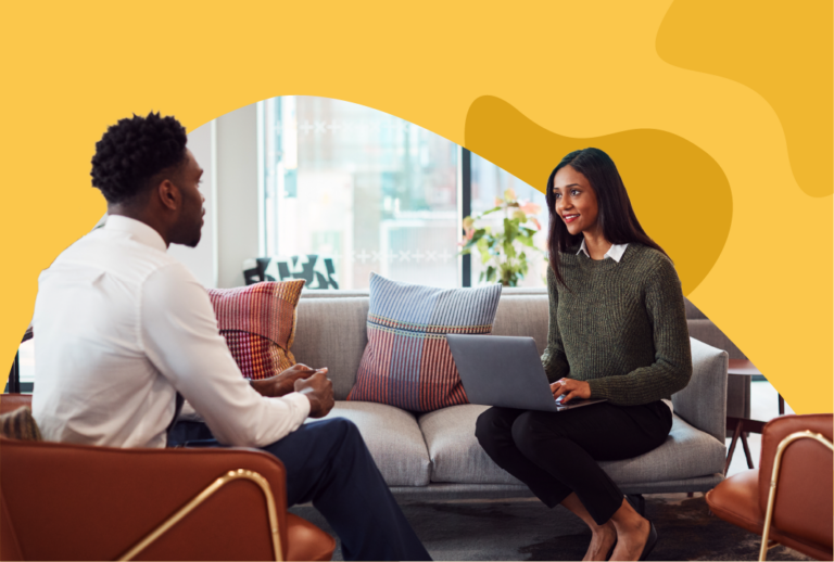 A manager sits with a laptop on a couch conducting a stay interview with an employee in a white collar shirt.