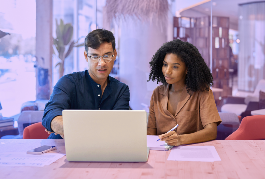 An employee in blue dress shirt is typing on laptop with a coworker in a brown shirt sitting next to them addressing bias in AI.