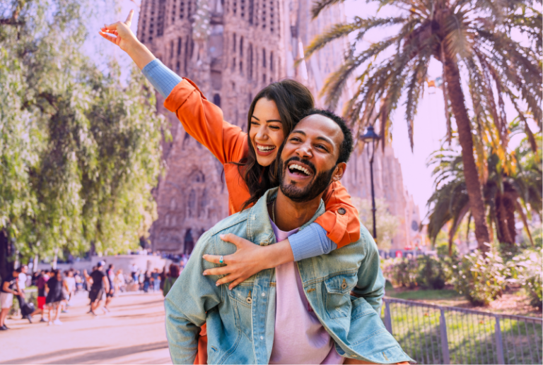 An employee is in Barcelona Spain, smiling, the employee's spouse is on the other's back, smiling and having fun.