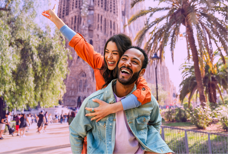 An employee is in Barcelona Spain, smiling, the employee's spouse is on the other's back, smiling and having fun.