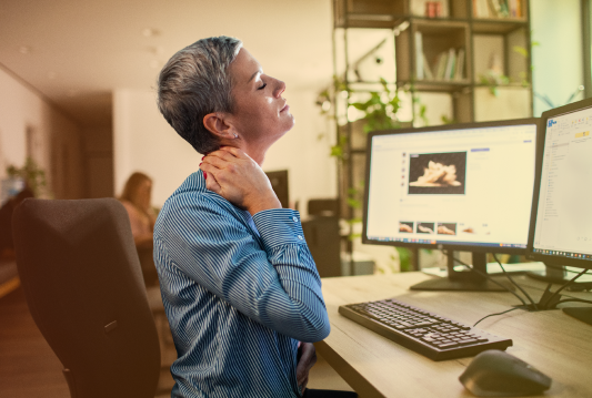 An employee is sitting at a desk in front of a computer monitor, rubbing their left shoulder.