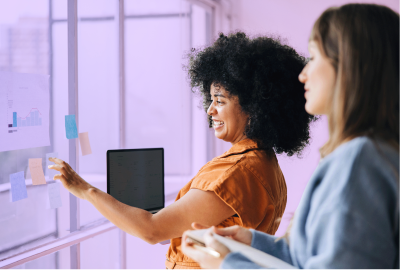 Two employees are standing in a boardroom by a whiteboard with post-it notes. They're making a plan based on HR trends.
