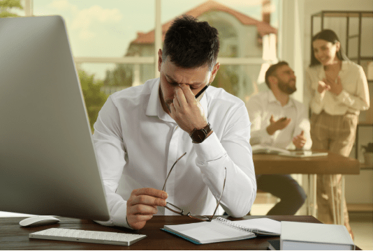 An employee appears to be the victim of workplace violence and harassment. They are sitting alone at their computer and look stressed. Two employees are talking to each other in the background.