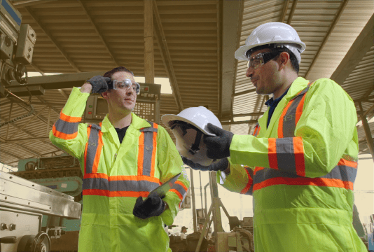 One worker is holding a hard hat and wearing PPE. They are looking at another employee who is not wearing all the PPE and forgot to wear his hard hat. They are in an industrial setting