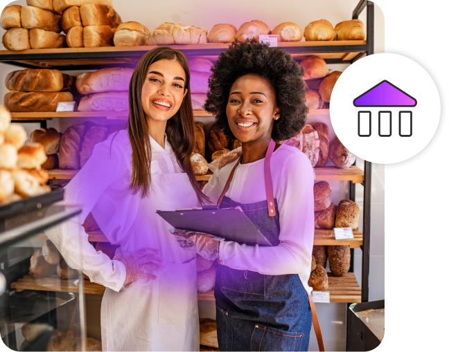 Two women smiling in a bakery wearing aprons. One of them is holding a clipboard.