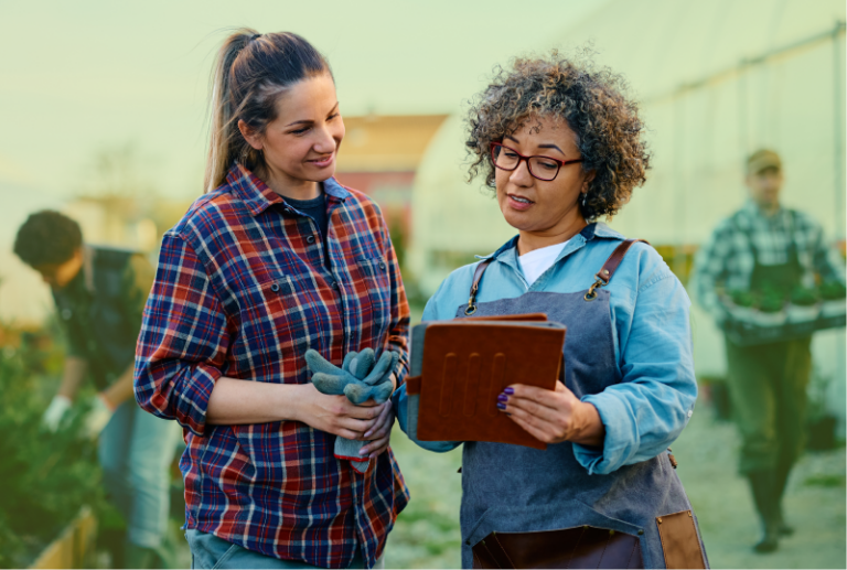 Two employees at a garden centre are standing beside each other looking at a tablet the one on the left is holding.
