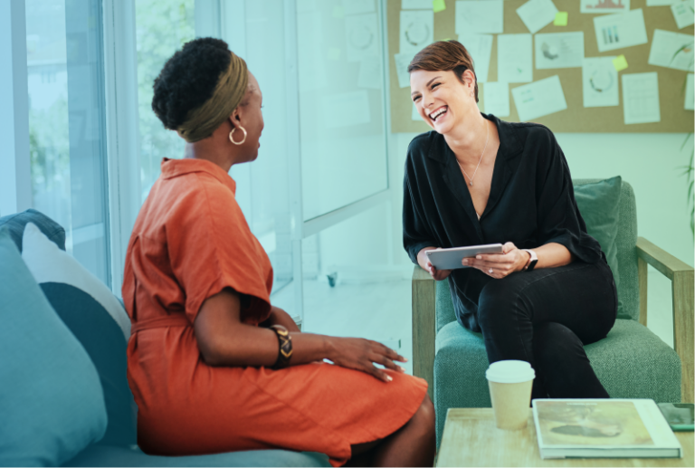 Two employees are sitting across a table from each other having a conversation. Both are smiling.