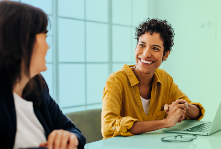 Two employees are sitting next to each other at a desk having a conversation.