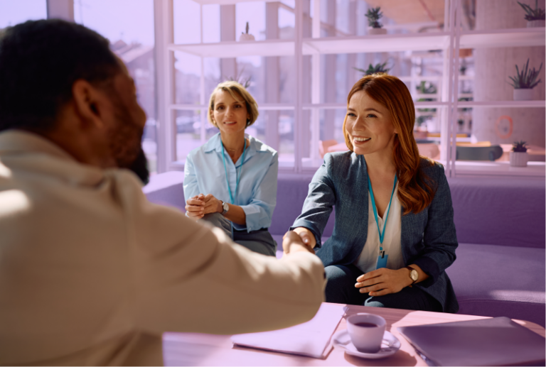 Two employees are using new hiring and recruitment strategies and are shaking a candidates hand across their desk while smiling.