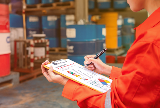 An employee in PPE is using a checklist to review WHMIS label requirements in a warehouse.