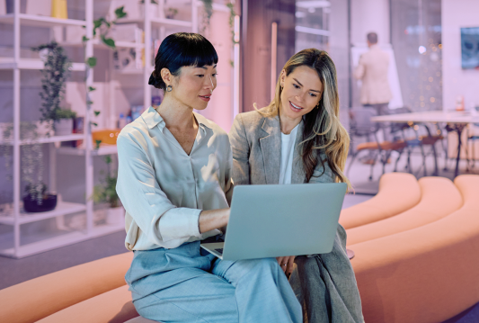 Two employees are standing beside each other, leaning on a table, looking at HR software on a laptop together.