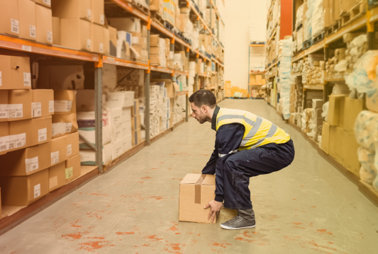 A warehouse worker is using proper lifting techniques to life a box from the floor of an aisle.