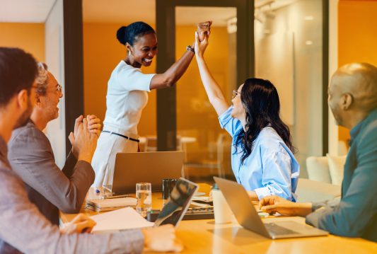 At a psychological health and safety training course, two employees are high-fiving each other across a table while four other employees cheer them on.